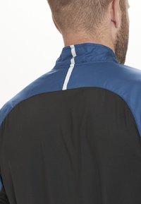 Schwarze Jacke mit blauem Kragen, auf der Rückseite mit einem vertikalen weißen Streifen. Glatte Textur, sportliches Design.
