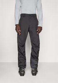 Pantalons de ski noirs imperméables avec une coupe droite, des poches zippées et des poignets renforcés. Portés par-dessus un haut de sport à manches longues gris et des chaussures de ski.