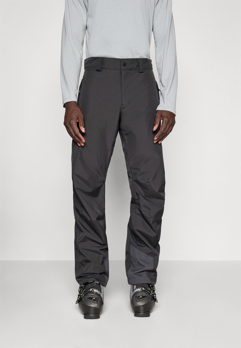 Pantalons de ski noirs imperméables avec une coupe droite, des poches zippées et des poignets renforcés. Portés par-dessus un haut de sport à manches longues gris et des chaussures de ski.