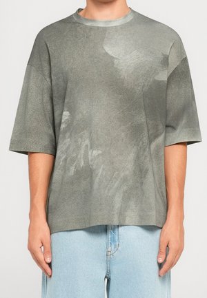 Person trägt ein lockeres, kurzärmliges grau-grünes T-Shirt mit Muster und hellblaue Jeans, steht mit entspannten Armen an den Seiten.