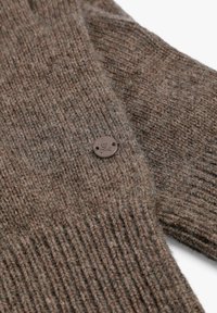 Detalle de suéter marrón tejido, con textura acanalada, un botón decorativo con un logo de calavera y un acabado suave y esponjoso.