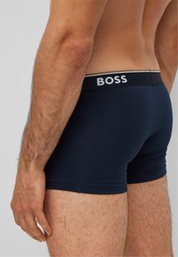 Schwarze Boxershorts mit einem grauen Bund, der das "BOSS"-Logo trägt. Der Stoff wirkt glatt und elastisch und schmiegt sich eng an den Körper.