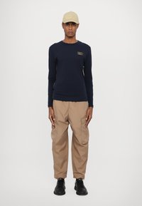 Tamsiai mėlyna ilgomis rankovėmis marškiniai su mažu auksiniu logotipu, smėlio spalvos kargo kelnės su tekstūruotu paviršiumi ir juodi sportiniai batai.