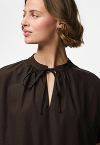 Tumši brūna blūze ar apaļu kakla izgriezumu, kurai ir sasienama detaļa un savilkta auduma daļa. Gluda tekstūra piešķir minimālistisku šarmu.