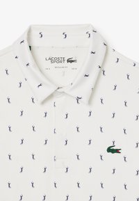 Valkoinen polo-paita, jossa on tavallinen mitoitus. Paidassa on tummansininen golfausteema ja rintataskussa kirjailtu vihreä krokotiililogo.