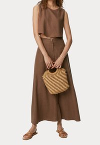 Top corto de lino marrón combinado con una larga falda marrón con detalles de botones. Sosteniendo un bolso de paja tejido. Sandalias marrones de tiras.