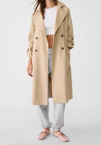 Vrouw in een beige trenchcoat over een witte crop top, een grijze joggingbroek en lichtroze balletschoenen, staand tegen een witte achtergrond.