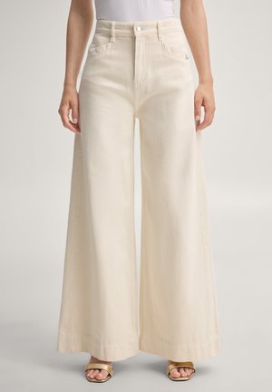 Persoon draagt een crèmekleurige high-waist jeans met wijde pijpen, een witte top en gouden open schoenen met hak, staand tegen een effen achtergrond.