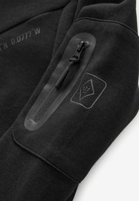 Sort sweatshirt med en lynlåslomme og et tekstureret logo-patch. Materialet er blødt med en glat, mat finish.