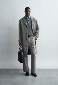 Sivi herringbone kaput, sivi džemper, plava košulja s kragnom, bež hlače, crni remen s rupicama, crna torba, crne cipele.