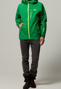 Grüne wasserdichte Jacke mit Kapuze, Reißverschluss vorne und Taschen. Mit gelben Akzenten und verstellbaren Bündchen. Getragen über dunkelgrauen Hosen und Stiefeln.
