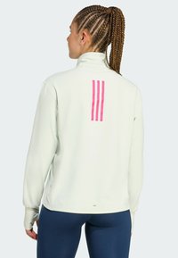 Kvinde med flettet hår iført en hvid bluse med lange ærmer og pink lodrette striber på ryggen samt navyblå leggings, stående sideways.
