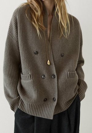 Kvinde iført oversized brun ribstrikket cardigan med sorte knapper, lommer, sorte bukser og en gylden vedhæng halskæde.