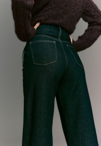 Dunkelblaue, hoch sitzende Jeans mit zwei Gesäßtaschen und kontrastierenden Nähten, kombiniert mit einem strukturierten, dunklen Strickpullover.