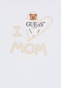 Bela majica s tekstom "I ❤️ MOM" v bež barvi, trikotno logotip z grafiko medveda in natisnjenim napisom "GUESS U.S.A." v črni barvi.