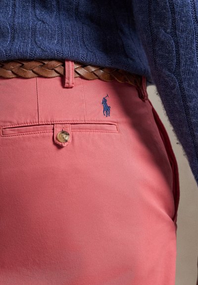 Nahaufnahme von korallenroten Hosen mit hinterer Knopftasche, geflochtenem braunem Ledergürtel und marineblauem Zopfmuster-Pullover.