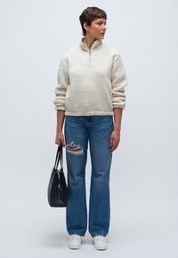 Svijetlo bež flis pulover s polu-zatvaračem na ovratniku, uparen s poderanim plavim trapericama i bijelim tenisicama, u ruci drži crnu torbicu.