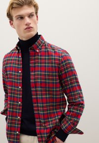 Camisa de cuadros rojos con cuello abotonado, con acentos en azul, verde y amarillo. Usada sobre un suéter de cuello alto negro, con mangas largas.