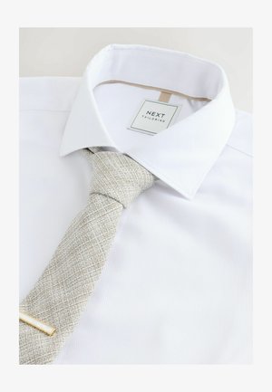 Bijela košulja sa strukturiranim ovratnikom i teksturiranom bež kravatom. Kravata ima dijagonalni uzorak i jednostavnu zlatnu kopču za kravatu.