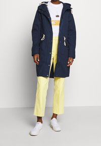 Mörkblå parka med dragsko i huvan, framknappar och fickor med dragkedja. Matchad med ljusgula manchesterbyxor och vita sneakers.