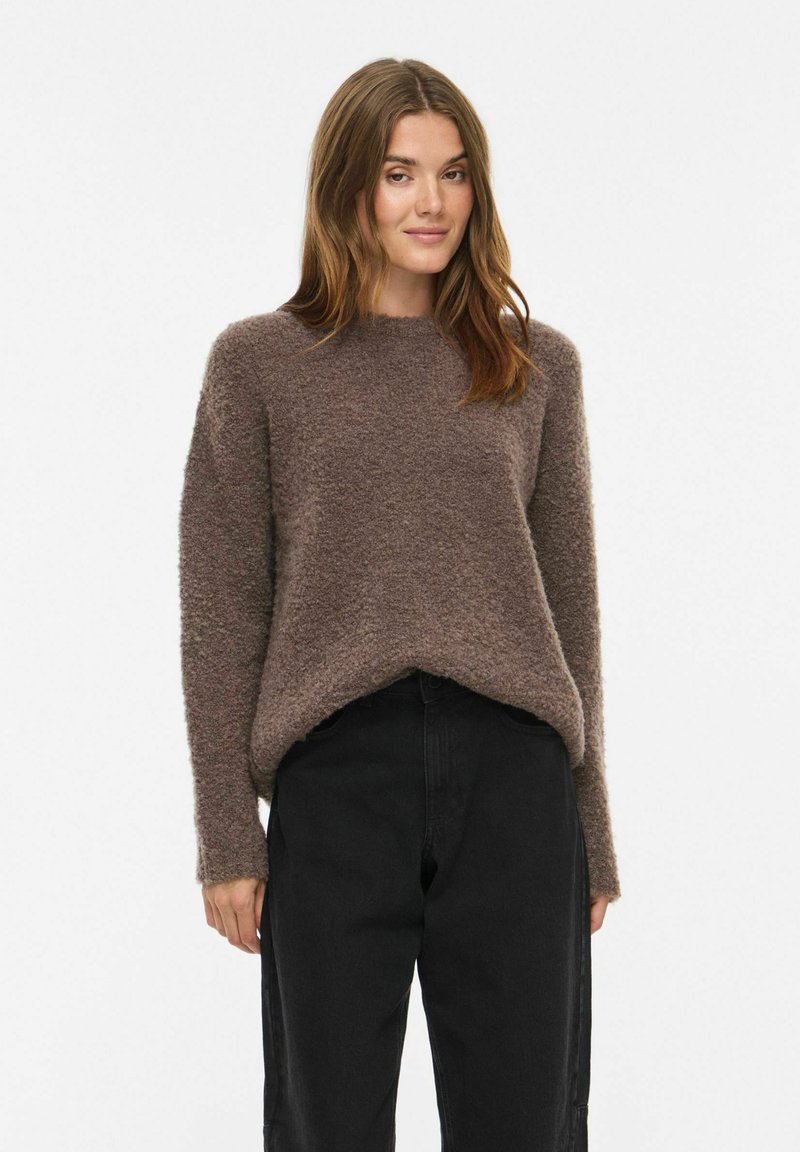 Femme portant un pull marron duveteux et un pantalon noir, debout avec un léger sourire sur un fond clair uni.