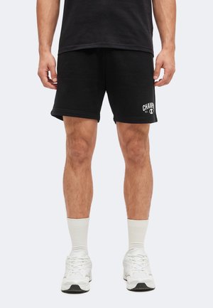 Mann trägt schwarze Champion-Shorts, weiße Sportsocken und weiße Sneaker, steht vor einem schlichten hellen Hintergrund.
