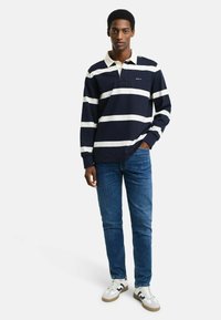 Polo de manga larga a rayas en azul marino y blanco, con cuello abotonado, combinado con jeans azules y zapatillas blancas con detalles en gris.