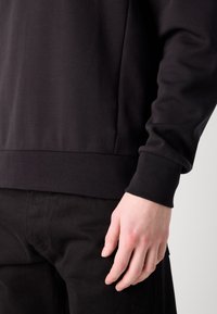 Main d'homme reposant à côté du corps, portant une chemise noire à manches longues avec un poignet côtelé et un pantalon noir sur un fond neutre.