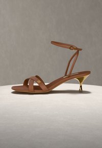 Καφέ δερμάτινο σανδάλι με λουράκια, χρυσό τακούνι kitten και αγκράφα στον αστράγαλο, απεικονίζεται σε φωτεινή επιφάνεια πάνω σε απλό γκρι φόντο.