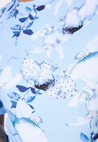 Camisa de manga corta azul claro con estampado floral; presenta patrones en azul y blanco y acentos de puntos; textura de tela suave y lisa.