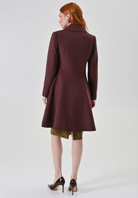 Cappotto in misto lana bordeaux con corpo aderente e gonna svasata, maniche lunghe e colletto classico. Indossato sopra un vestito oliva, abbinato a tacchi.