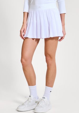 Kobieta w białej plisowanej spódnicy, białej bluzce z długim rękawem, białych skarpetkach i białych trampkach stojąca na tle jednolitego tła.