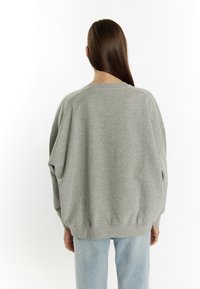 Felpa grigia oversize con maniche lunghe, colletto e vita a costine; indossata con jeans azzurri chiari. Texture liscia, design informale.