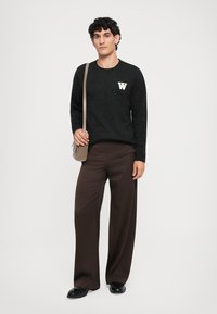 Svart strikket genser med hvit "W" logo, parret med brune vide bukser og svarte skinnsko. Crossover-veske i lys brun.