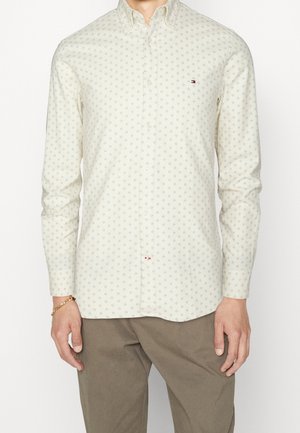 Hombre con una camisa de botones de color crema claro con un pequeño patrón geométrico azul y pantalones marrones, de pie contra un fondo blanco.