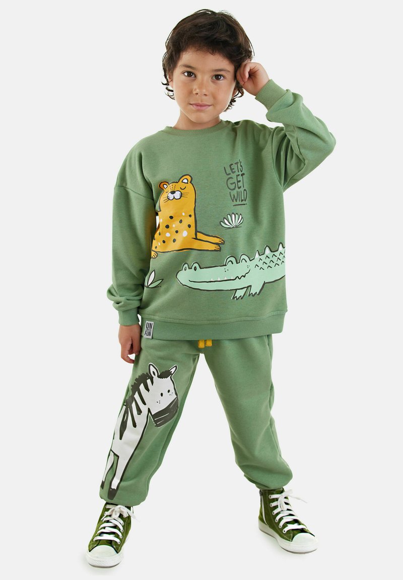 Dreng i grøn sweatshirt og bukser med tegneserieprint af leopard, krokodille og zebra, poserende med hånden nær hovedet og grønne høj-top sneakers.