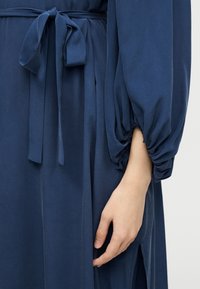 Χέρι που ακουμπά δίπλα σε μπλε σκούρο φόρεμα με ζώνη στη μέση και με φουσκωτό μανίκι με πιέτες, πάνω σε απλό φόντο.