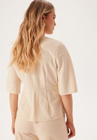 Beige kortærmet top med tætsiddende talje og plisseringer bagpå, lavet af struktureret stof, glat finish og buet halsudskæring.