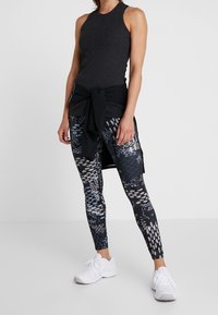 Kolcharcoal ribbad linne, svart knuten midjelager, mönstrade leggings med blå, grå och vita mönster, och vita träningsskor.