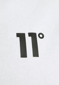 "11°" imprimé en noir sur un tissu blanc texturé, probablement une partie d'un vêtement ou d'un matériau textile.