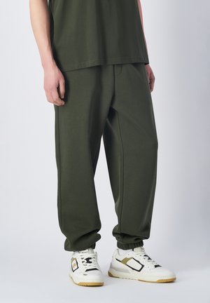 Pantalones deportivos de color verde oliva con puños elásticos, corte relajado y bolsillos laterales. Combinados con zapatillas blancas que tienen detalles en negro y dorado.