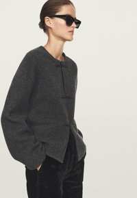 Giacca di lana grigia con colletto rotondo, maniche oversize e due chiusure a pulsante. Abbinata a pantaloni neri strutturati.