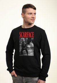 Schwarzer Sweatshirt mit einem großen Grafik eines schwarz-weißen Szenes aus "Scarface" und fett gedrucktem roten Text darüber mit der Aufschrift "SCARFACE."