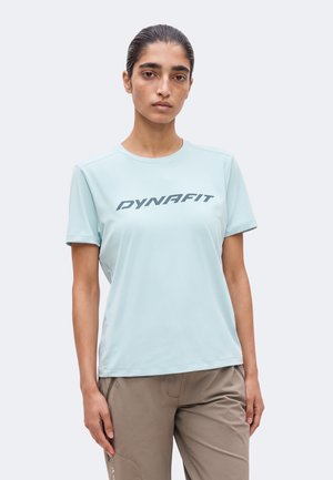 Persona joven con el pelo peinado hacia atrás, con una camiseta Dynafit azul clara y pantalones topo, de pie frente a un fondo sencillo.