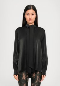 Black silk blouse with a tie neck, long sleeves, and a relaxed fit. Smooth texture with subtle sheen. Paired with patterned leggings.