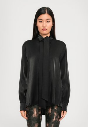 Black silk blouse with a tie neck, long sleeves, and a relaxed fit. Smooth texture with subtle sheen. Paired with patterned leggings.