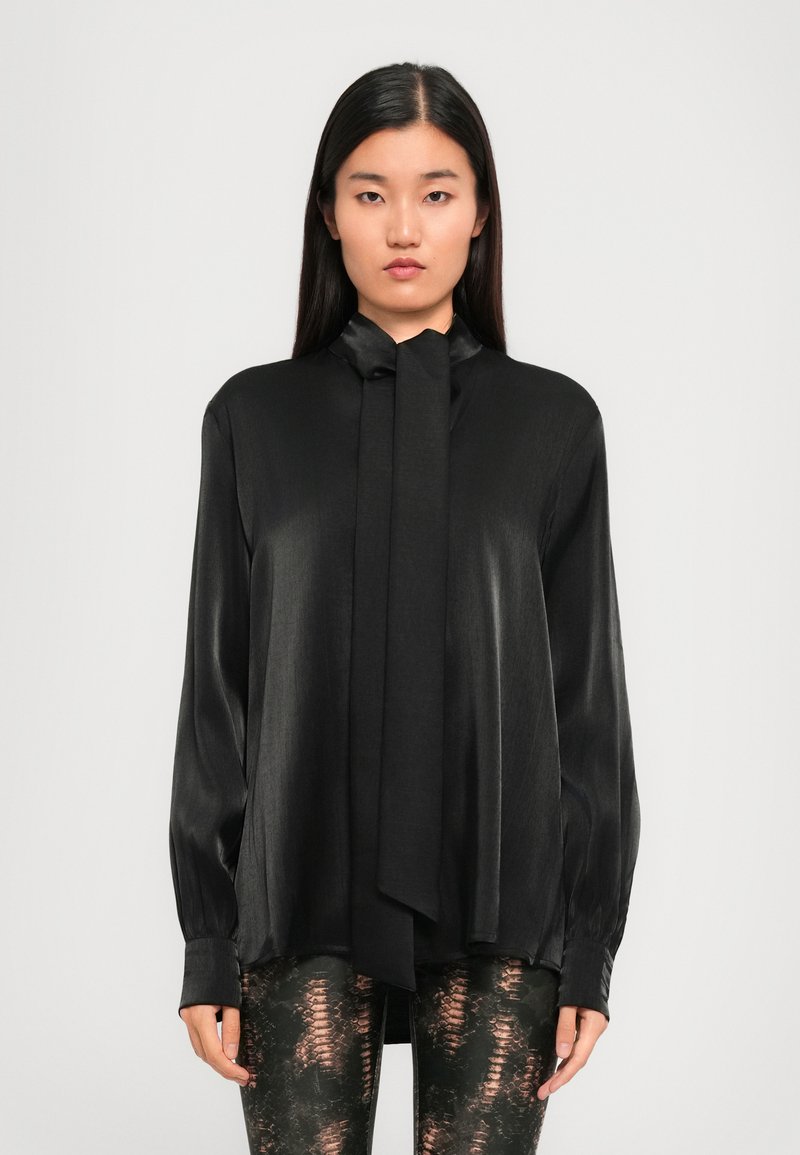 Blusa di seta nera con colletto a legaccio, maniche lunghe e vestibilità rilassata. Texture liscia con una leggera lucentezza. Abbinata a leggings fantasia.