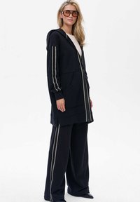 Czarna kurtka z kapturem, z srebrnymi paskami po bokach, kieszeniami z przodu i zamkiem błyskawicznym, zestawiona z luźnymi czarnymi spodniami. Teksturowany materiał.