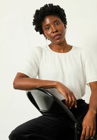 Weißes, strukturiertes Blusenshirt mit kurzen Ärmeln und dezenten, glänzenden Akzenten, kombiniert mit schwarzen Hosen, vor einem neutralen Hintergrund.