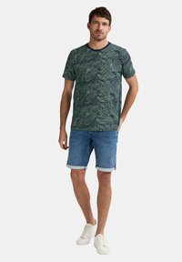 Herreskjorte med korte ærmer i et bladmønster i grøn og marineblå, kombineret med blå denimshorts og hvide sneakers. Enkel, afslappet design.
